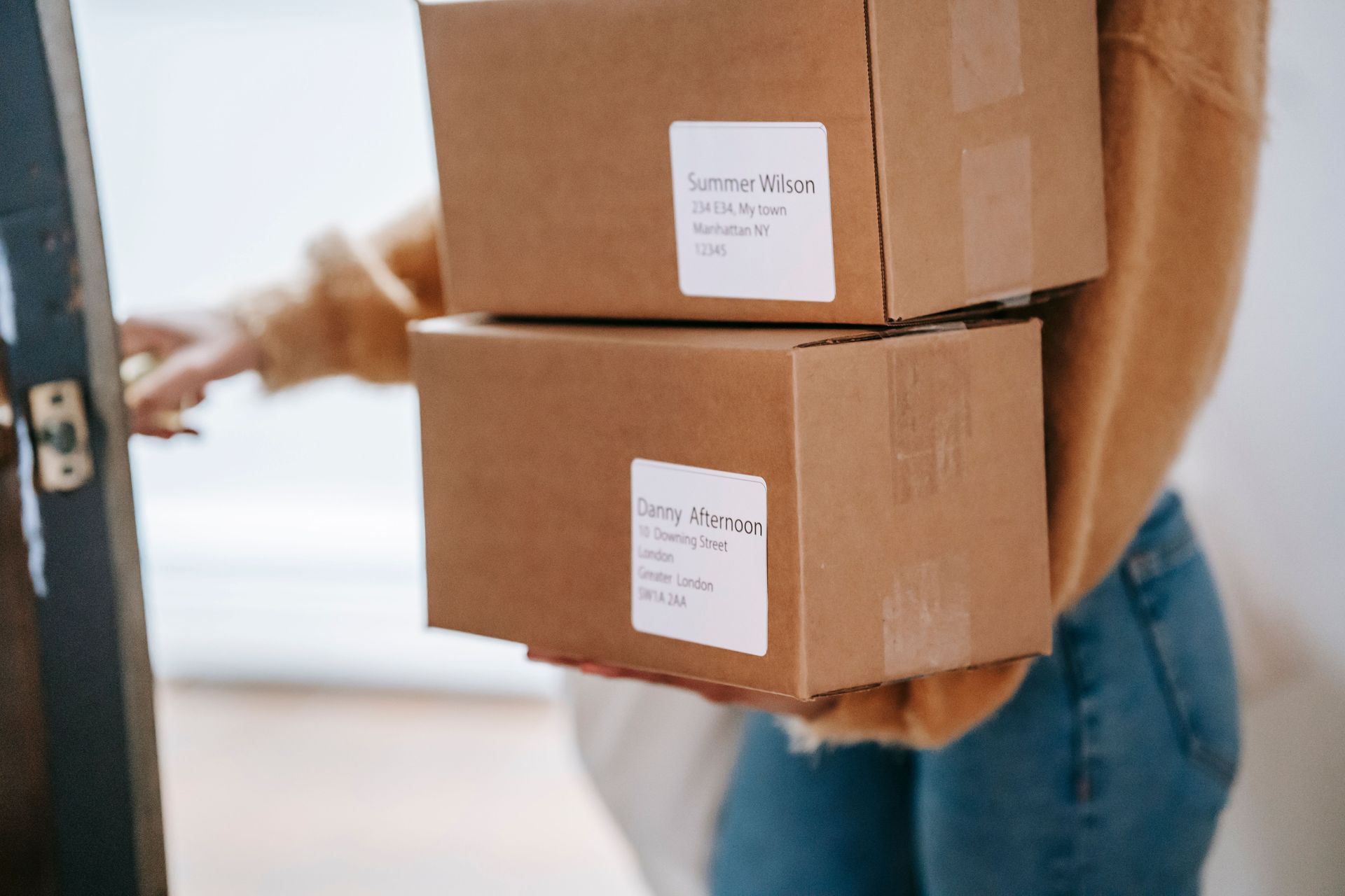 Crop anonymous female wearing casual outfit carrying carton parcels with address labels opening door of post office