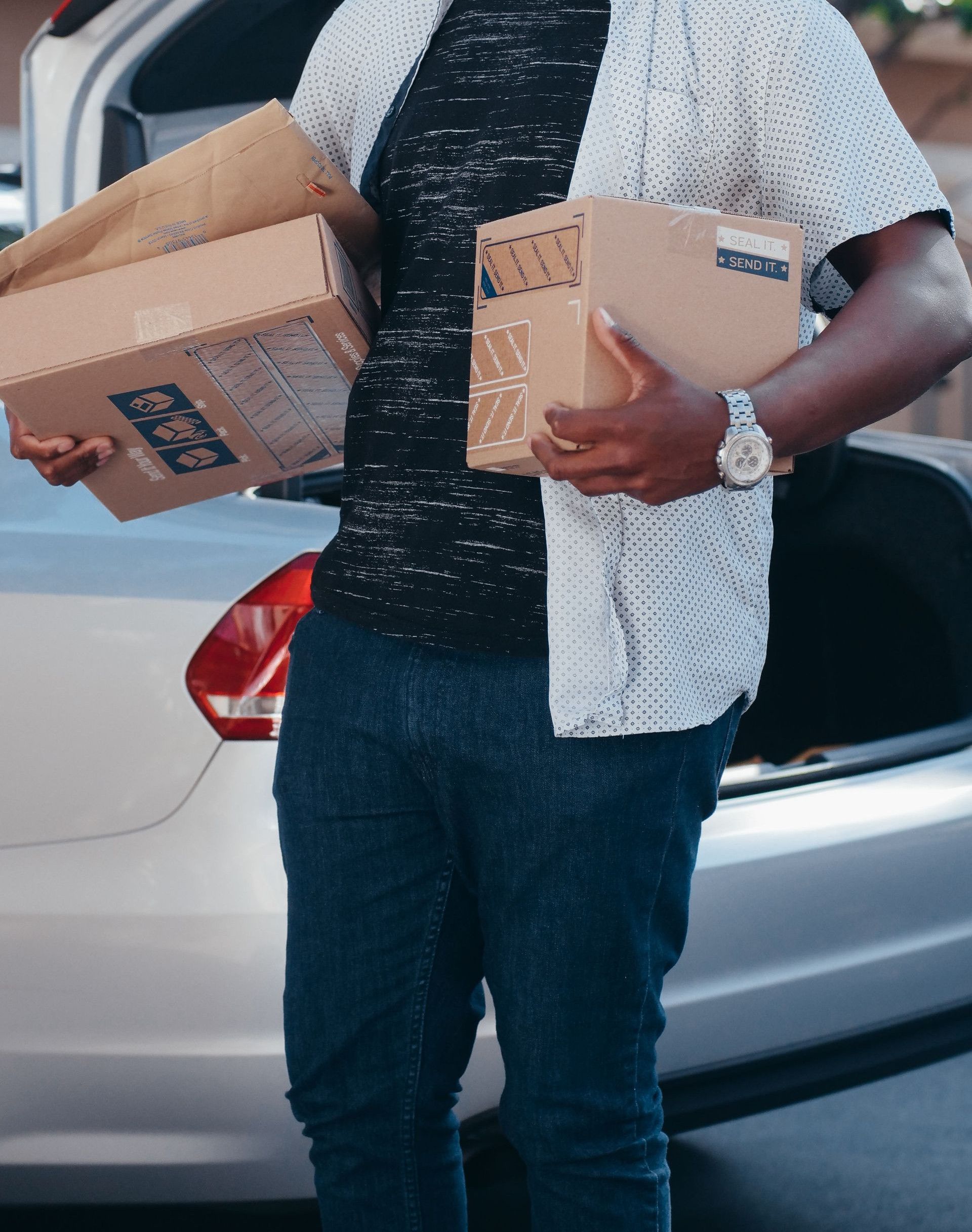 A cheerful man holding packages stands by an open car trunk outdoors, ideal for delivery and logistics concepts.