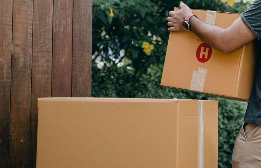 Side view of serious young ethnic man in casual clothes unloading cardboard boxes out of trunk of modern car in blooming green backyard while moving in new house